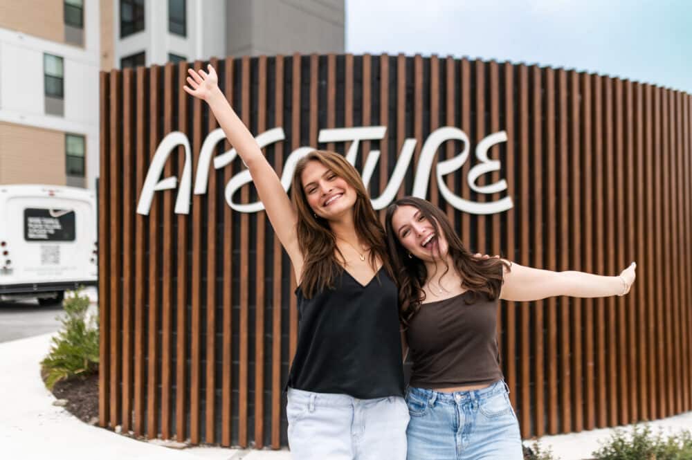 Two young women smiling and posing joyfully in front of a modern wooden slat wall with a large white "Aperture" sign. One woman has her arm raised high in the air while the other leans in close.