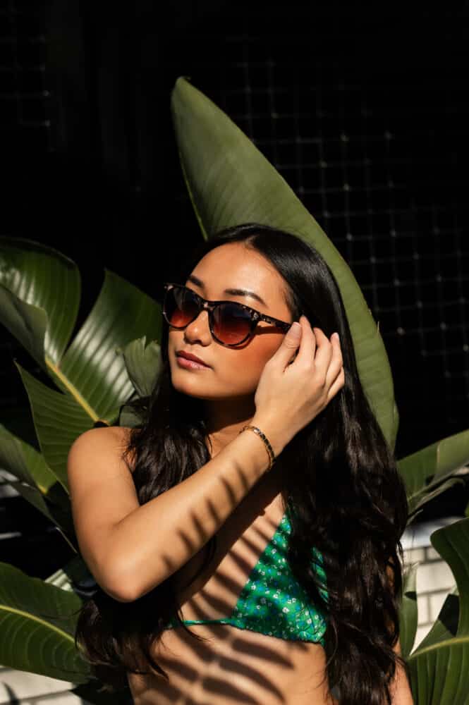 A close-up portrait of a young woman wearing sunglasses and a green floral bikini top, posing in front of large green tropical leaves. Shadows from the leaves create a striped pattern across her face and shoulder.