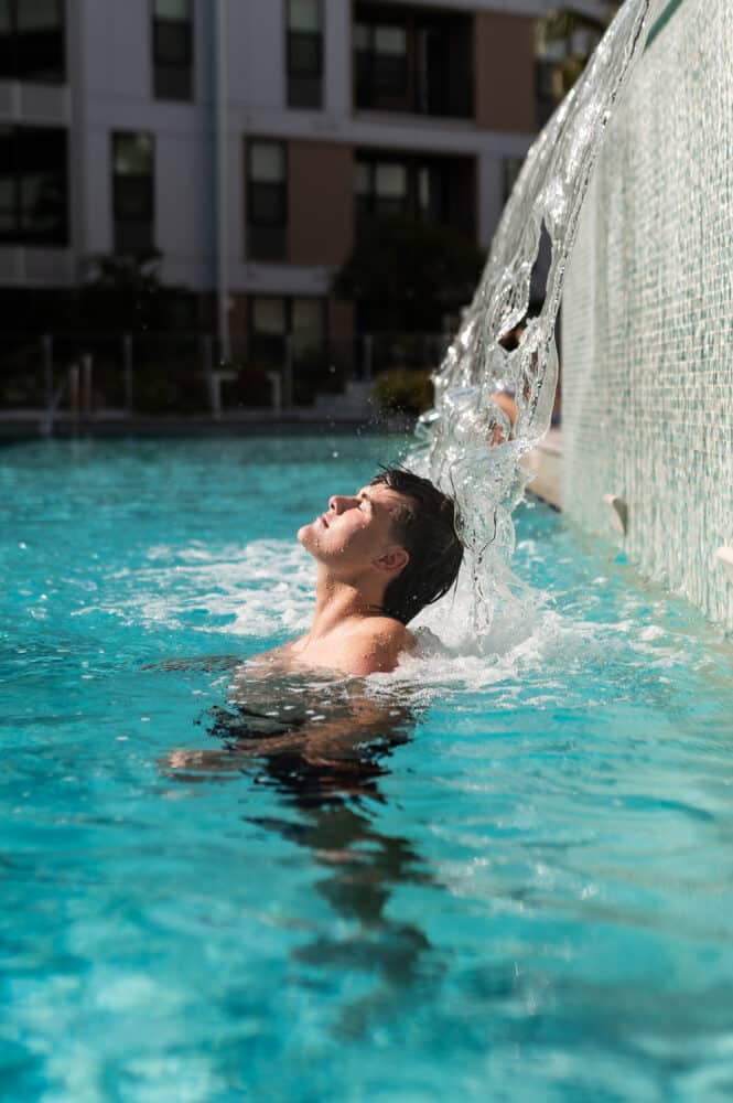 A young man submerged in a swimming pool, leaning his head back directly under a decorative waterfall feature. The water splashes over his face as he enjoys the refreshing flow.