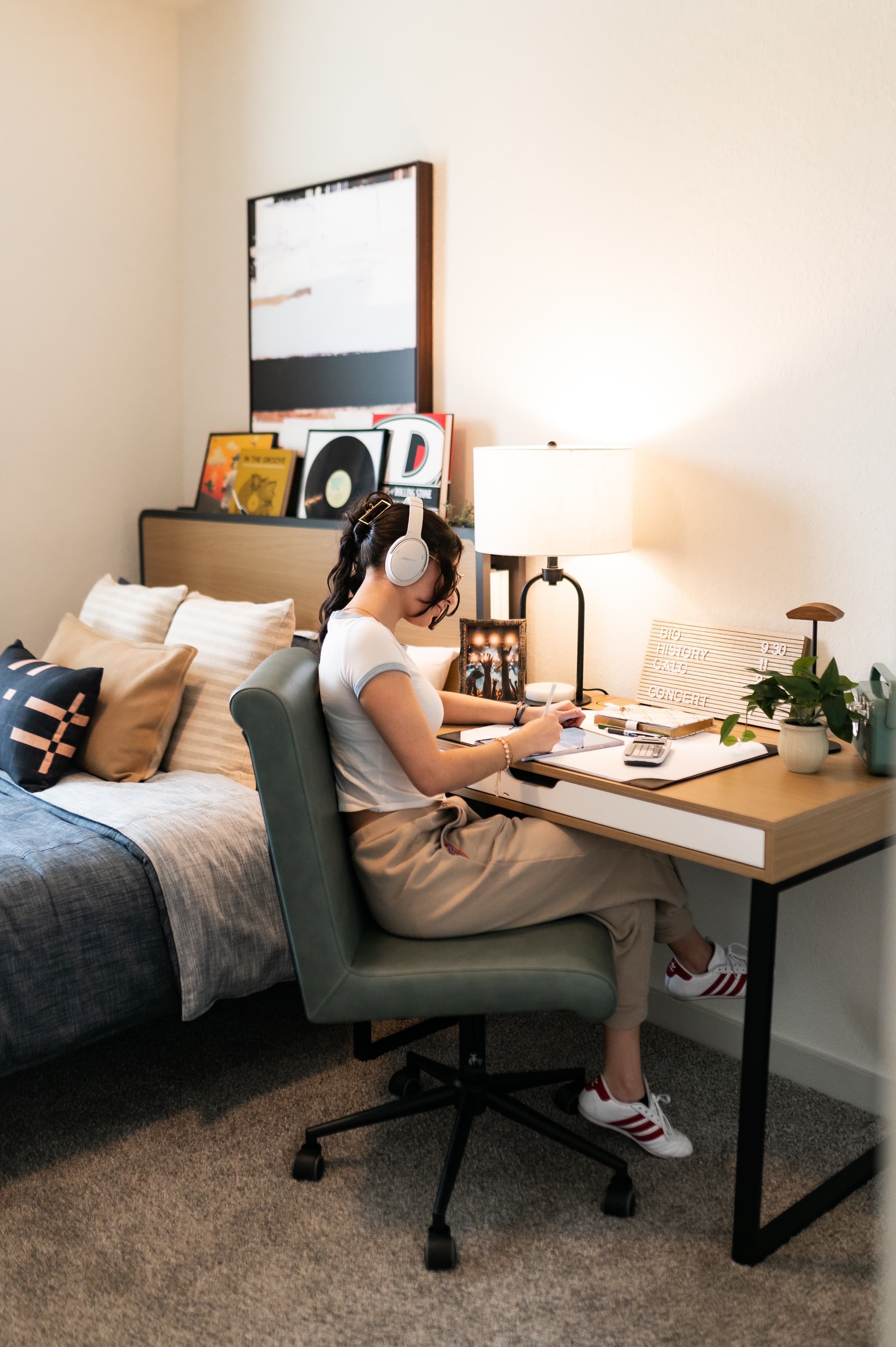 A young woman sitting at a modern wooden desk in a bedroom, wearing white headphones and focused on her studies. The room is decorated with a stylish bed, a warm desk lamp, and vinyl records on the headboard.