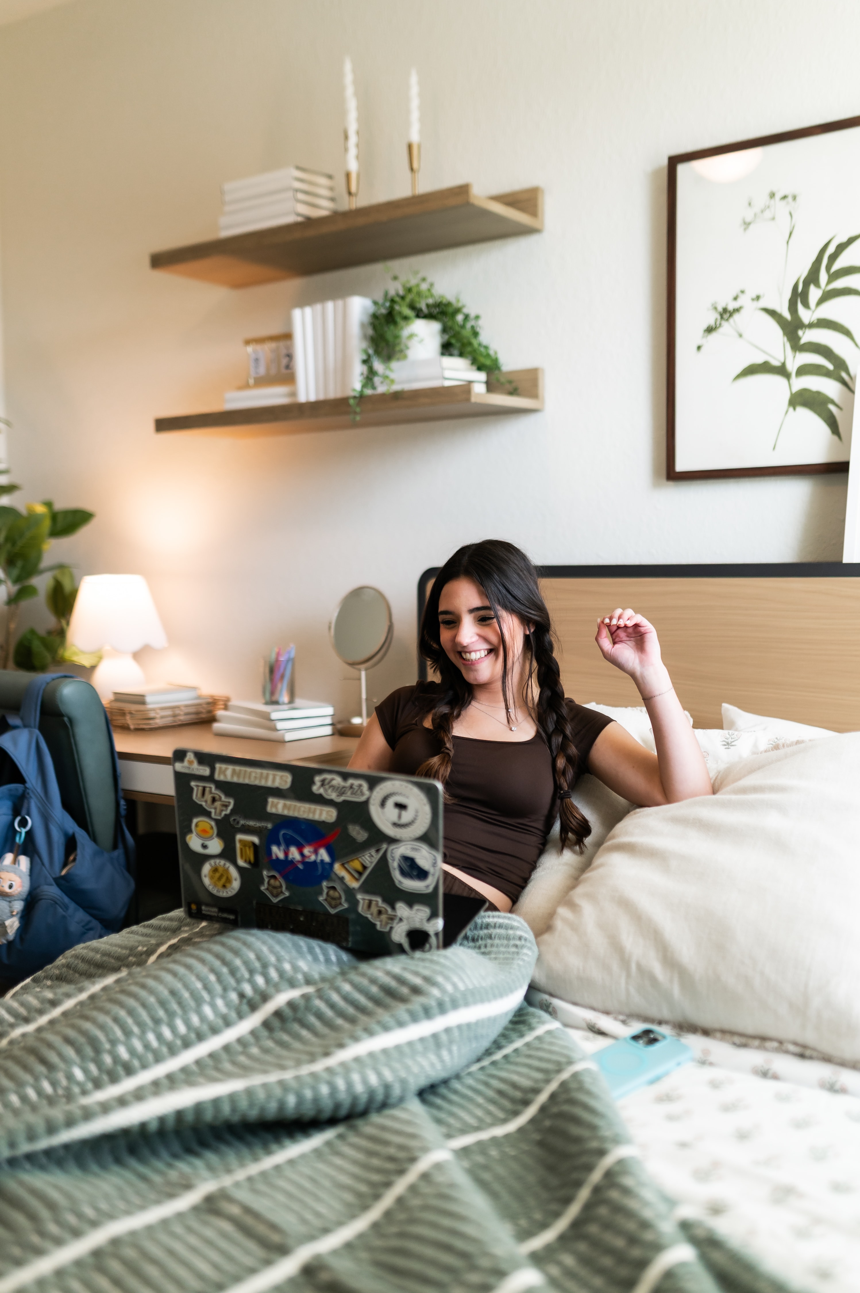 A young woman with braided hair sitting in bed, leaning against a neutral-toned headboard while looking at a laptop covered in colorful stickers. She is laughing and appears to be on a video call.