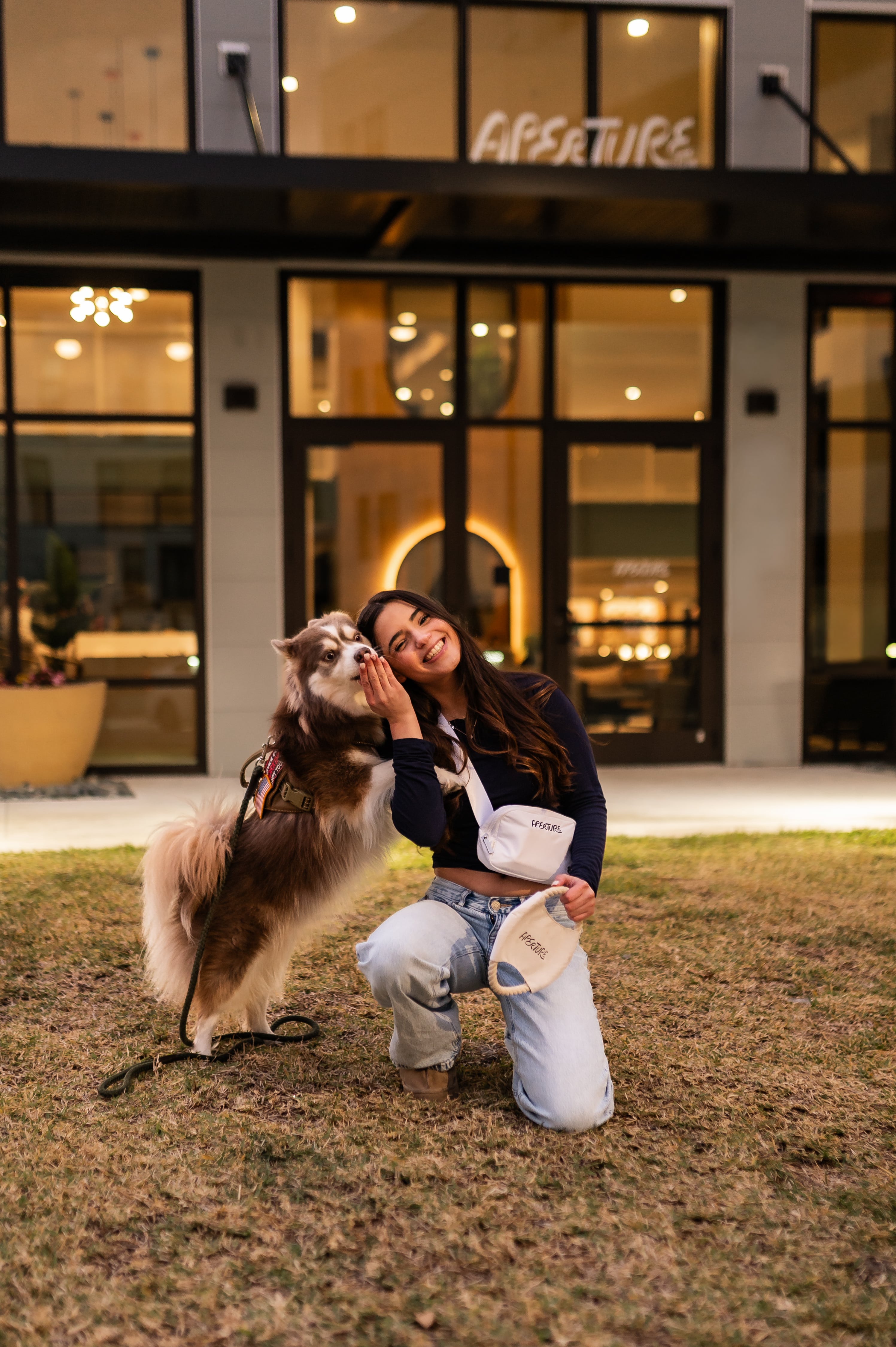 Two young women standing side-by-side in front of a bright, backlit bathroom mirror. One is applying lip liner while the other holds a makeup product, both focused on their reflections as they get ready.